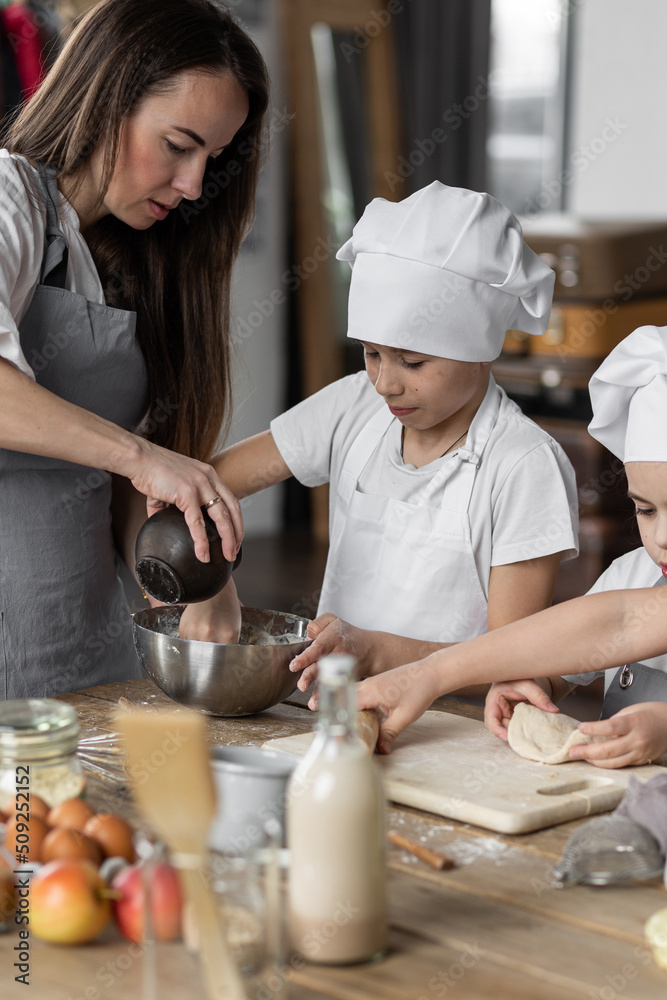 Mother and her little cute kids baking homemade sweet pie together, having fun. Home bakery, little kids in process of food preparation in the kitchen at home, helping mother, doing chores Mother and her little cute kids baking homemade sweet pie together, having fun. Home bakery, little kids in process of food preparation in the kitchen at home, helping mother, doing chores
