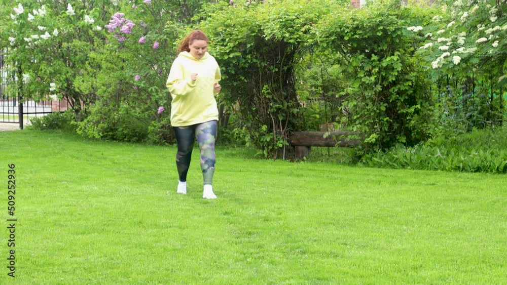 Red-haired curly plus size fat woman with excess weight in yellow sport hoodie doing cardio fitness, running and jogging in green natural park, meadow. Lose weight, burn calories. Prepare to summer Red-haired curly plus size fat woman with excess weight in yellow sport hoodie doing cardio fitness, running and jogging in green natural park, meadow. Lose weight, burn calories. Prepare to summer