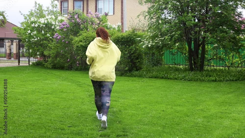 Red-haired curly plus size fat woman with excess weight in yellow hoodie doing cardio fitness, running fast in the green backyard outdoors. Nature, park and house on background. Lose weight, rearview Red-haired curly plus size fat woman with excess weight in yellow hoodie doing cardio fitness, running fast in the green backyard outdoors. Nature, park and house on background. Lose weight, rearview