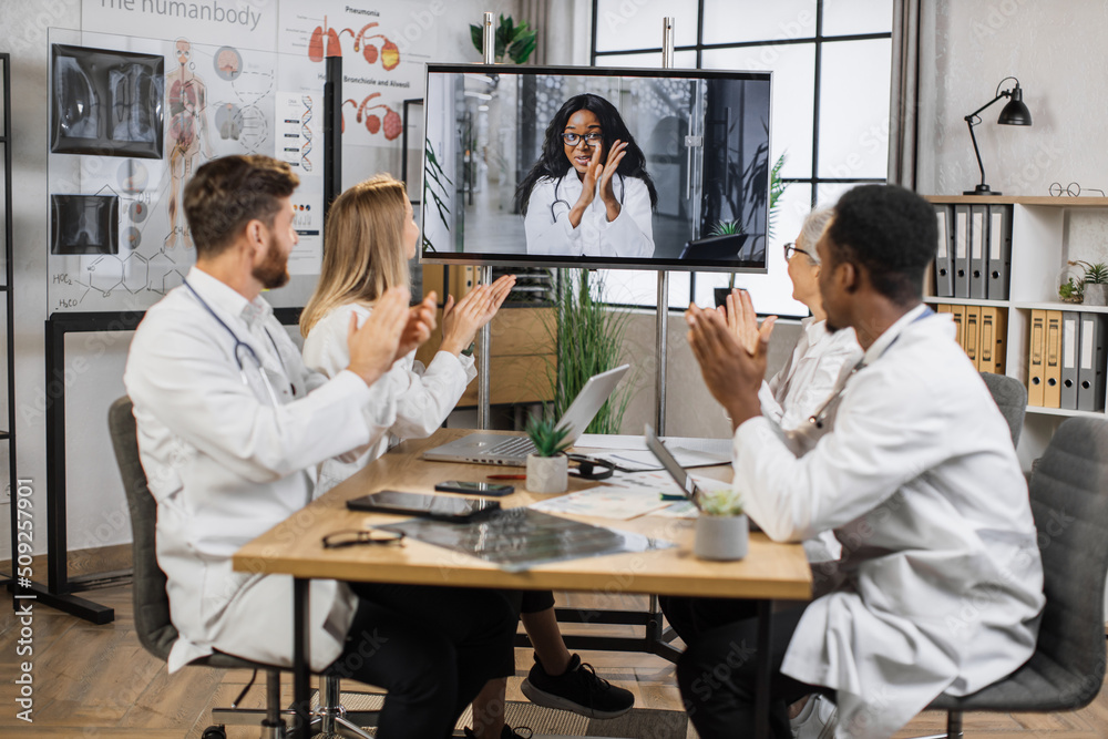 Team of multicultural doctors in white lab coats clapping hands during online video conference after successful speech of excited female scientist. Focus on african american woman on monitor screen. Team of multicultural doctors in white lab coats clapping hands during online video conference after successful speech of excited female scientist. Focus on african american woman on monitor screen.