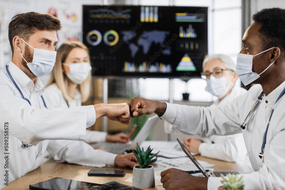 Obraz premium Team of male physicians in face masks giving fist bump while sitting at clinic table with female coworkers on background. Preventive multicultural colleagues making deal during coronavirus outbreak.