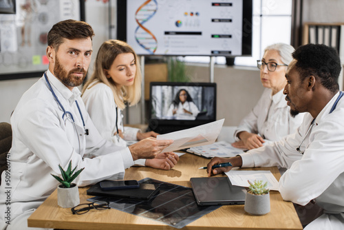 Caucasian male doctor looking at camera while sitting at desk with presentation list in hands. Multiracial coworkers sitting near and listening african american woman during video call on laptop.