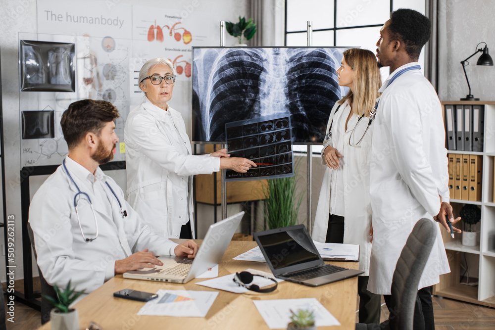 Obraz premium Caucasian senior therapist smiling and looking at camera while pointing on MRI scan in boardroom. Group of multiethnic colleagues in white lab coat discussing clinical case for making right diagnosis.