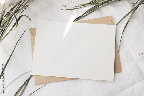 Summer wedding stationery mock-up  desk scene. Blank horizontal greeting card  dry green palm leaf. In sunlight. White linen table background. Tropical styled photo  web banner. Flat lay  top view.