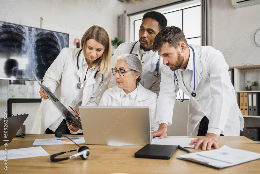 Multiethnic physicians discussing x ray scan while standing around senior female doctor in hospital. Competent elder woman sitting at desk in front of laptop and giving consultation for collegues. Multiethnic physicians discussing x ray scan while standing around senior female doctor in hospital. Competent elder woman sitting at desk in front of laptop and giving consultation for collegues.