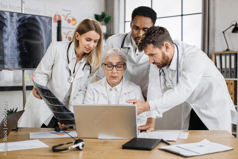 Multiethnic physicians discussing x ray scan while standing around senior female doctor in hospital. Competent elder woman sitting at desk in front of laptop and giving consultation for collegues. Multiethnic physicians discussing x ray scan while standing around senior female doctor in hospital. Competent elder woman sitting at desk in front of laptop and giving consultation for collegues.