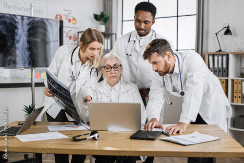 International team of three interns having urgent meeting with senior female doctor in hospital conference room. Workerks in white lab coats using modern laptop for analysing patients medical record. photo