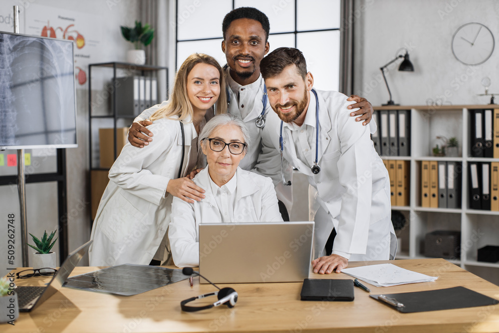 Obraz premium Group of multicultural medical workers in white lab coats hugging and looking at camera with smile in bright clinic boardroom. Effective teamwork and healthcare cooperation concept.