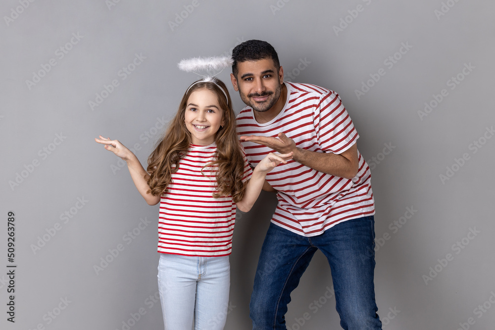 Portrait of smiling happy man showing her charming daughter with with halo over head, cute little girl raised arms, family looking at camera. Indoor studio shot isolated on gray background. Portrait of smiling happy man showing her charming daughter with with halo over head, cute little girl raised arms, family looking at camera. Indoor studio shot isolated on gray background.