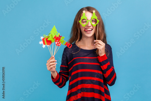 Funny positive woman wearing striped casual style sweater, holds Christmas party props, covers eyes with paper glasses, showing tongue out, festive mood. Indoor studio shot isolated on blue background