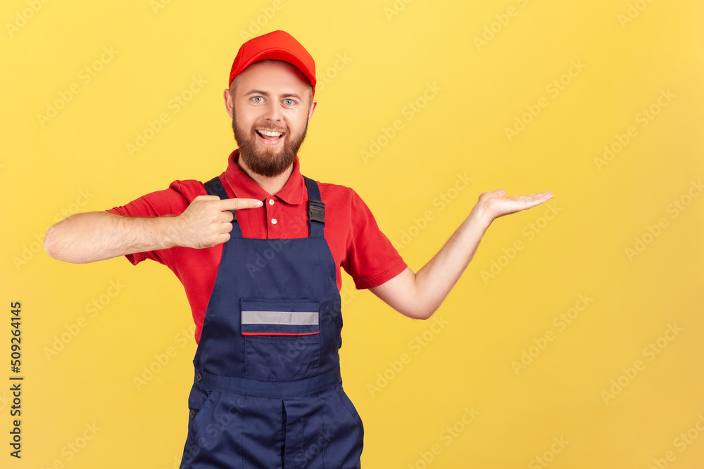 Smiling optimistic worker man standing with positive expression, pointing finger aside, presenting copyspace on his palm, advertise his service. Indoor studio shot isolated on yellow background.