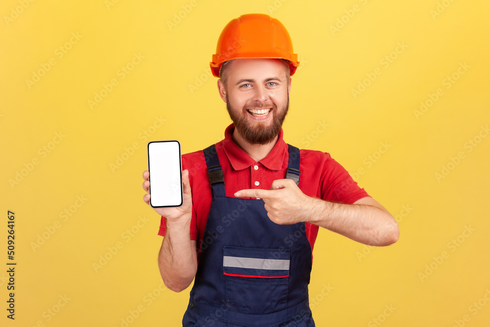 Satisfied builder man or repairman in uniform and protective helmet showing cell phone with blank empty screen with copy space, advertise his service. Indoor studio shot isolated on yellow background. Satisfied builder man or repairman in uniform and protective helmet showing cell phone with blank empty screen with copy space, advertise his service. Indoor studio shot isolated on yellow background.