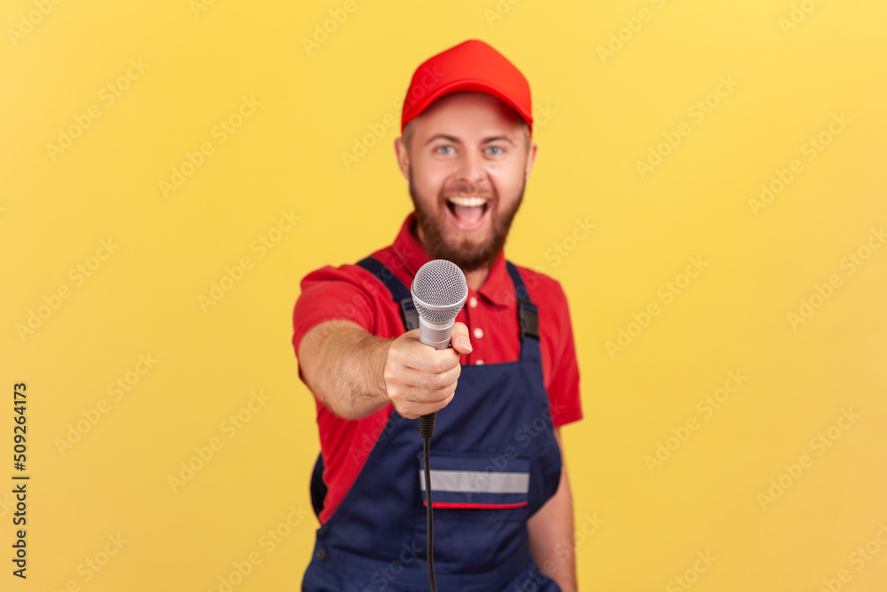 Portrait of excited bearded worker man in blue uniform and cap standing and offering microphone looking at camera with happy facial expression. Indoor studio shot isolated on yellow background. Portrait of excited bearded worker man in blue uniform and cap standing and offering microphone looking at camera with happy facial expression. Indoor studio shot isolated on yellow background.