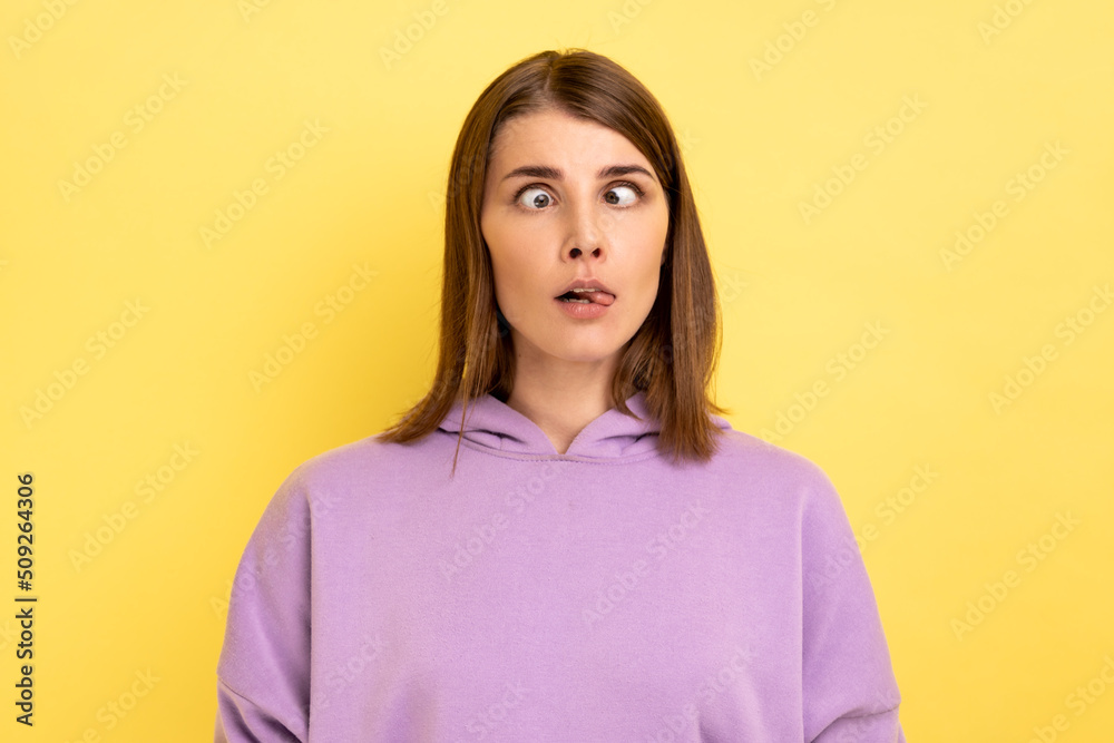 Portrait of young adult foolish woman with hair standing with crossed eyes and tongue out, childish behavior, wearing purple hoodie. Indoor studio shot isolated on yellow background. Portrait of young adult foolish woman with hair standing with crossed eyes and tongue out, childish behavior, wearing purple hoodie. Indoor studio shot isolated on yellow background.