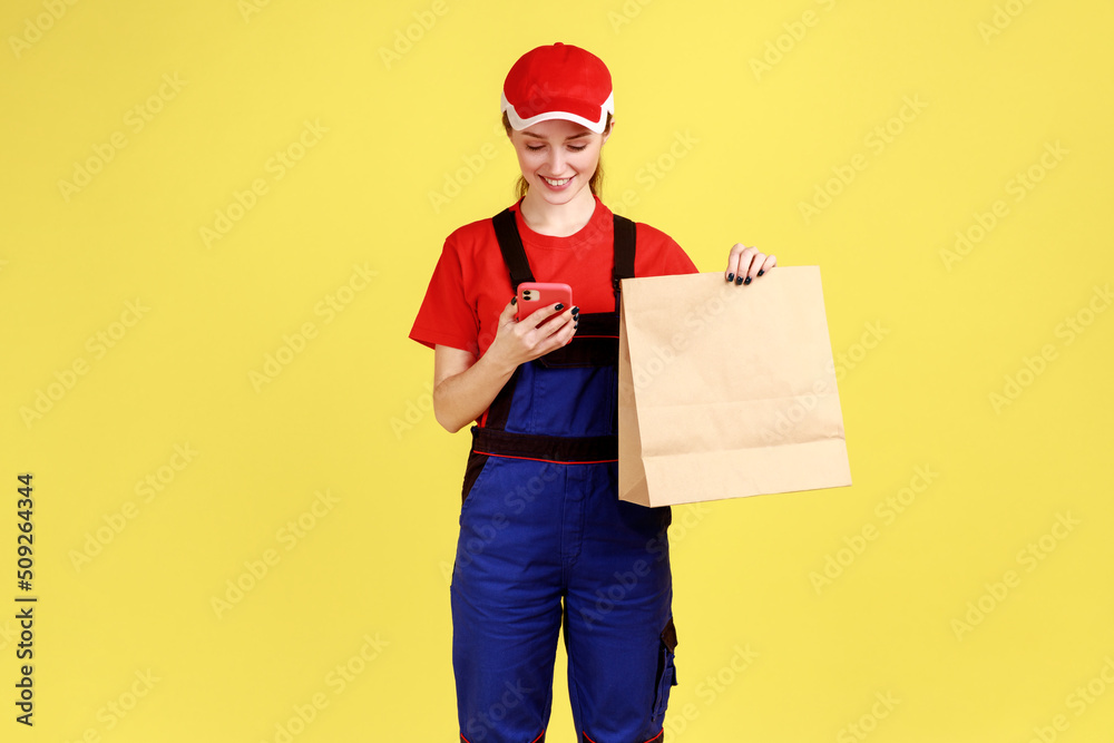 Portrait of smiling courier woman standing with paper parcel in hands, using mobile phone for communication with client, wearing overalls and red cap. Indoor studio shot isolated on yellow background. Portrait of smiling courier woman standing with paper parcel in hands, using mobile phone for communication with client, wearing overalls and red cap. Indoor studio shot isolated on yellow background.