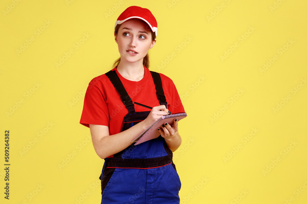 Portrait of thoughtful worker woman writing in paper notebook, writes down orders, looking away with pensive expression, wearing overalls and red cap. Indoor studio shot isolated on yellow background.