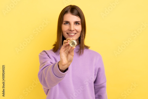 Look at that. Surprised excited woman standing holding bitcoin, paying attention at new digital cryptocurrency, wearing purple hoodie. Indoor studio shot isolated on yellow background.
