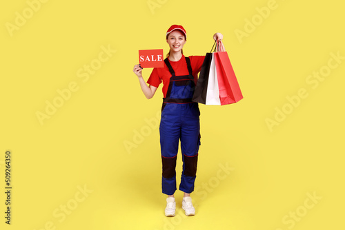 Full length portrait of delivery woman standing with shopping bags in hands, and showing card with sale inscription, wearing overalls and red cap. Indoor studio shot isolated on yellow background.