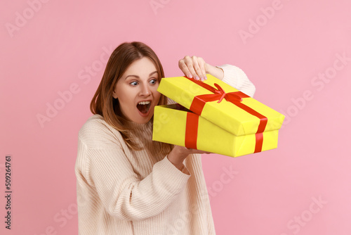Portrait of excited young adult blond woman looking inside yellow present box, being interested what inside, wearing white sweater. Indoor studio shot isolated on pink background. © khosrork