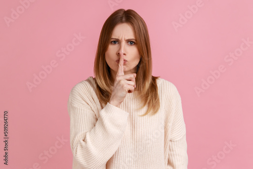 Portrait of blond woman keeping finger on lips making silence gesture, shushing asking to be quiet, secrecy concept, wearing white sweater. Indoor studio shot isolated on pink background.