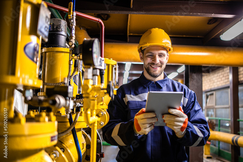 Power plant engineer in safety work wear standing by natural gas installations and pipeline checking consumption on tablet computer application.