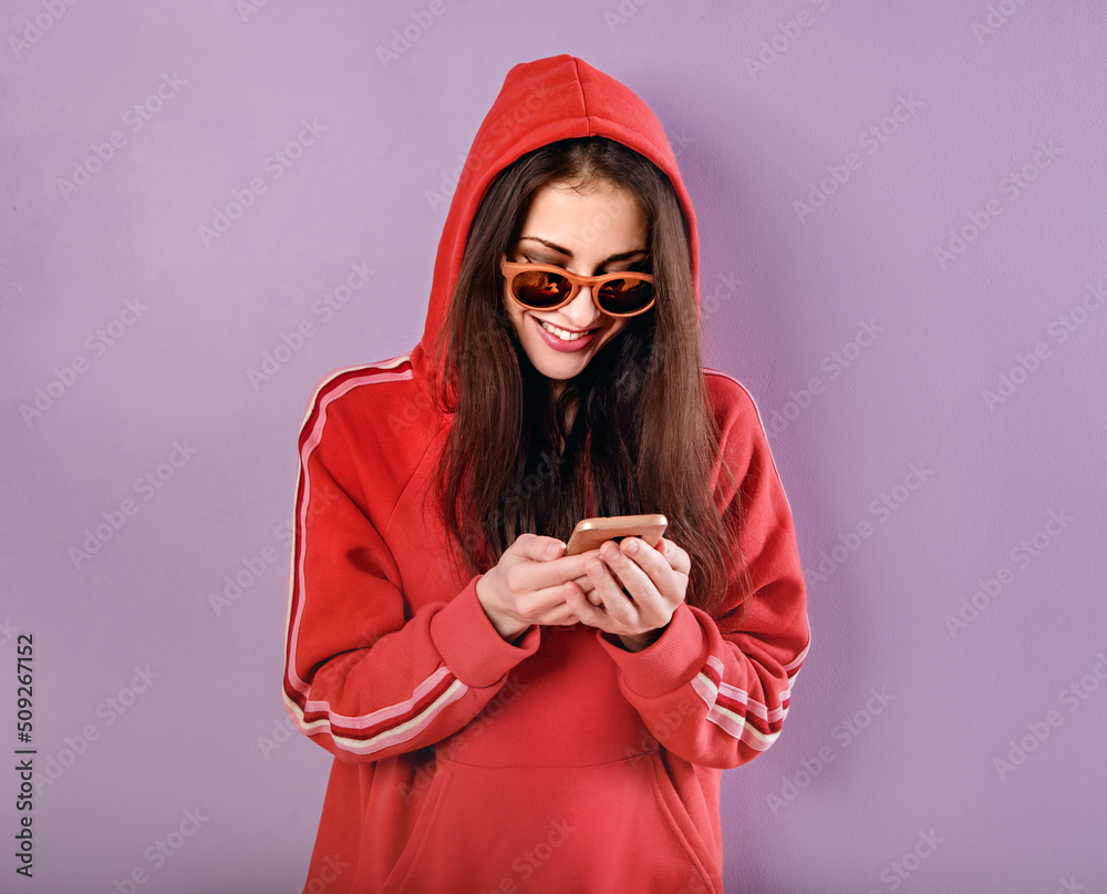Happy smiling brunette woman of caucasian holding in hand and use mobile cell phone in fashion red hoodie and sunglasses on purple bright background with empty copy space. Studio Happy smiling brunette woman of caucasian holding in hand and use mobile cell phone in fashion red hoodie and sunglasses on purple bright background with empty copy space. Studio