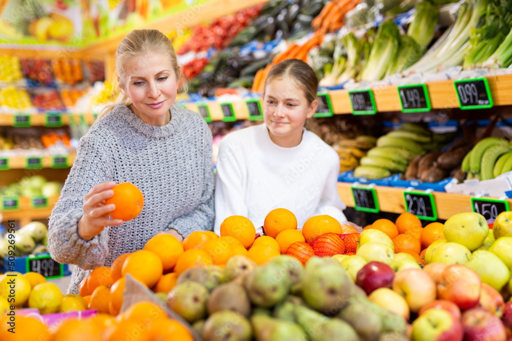 Cheerful teen girl with mother choosing sweet ripe oranges while shopping in greengrocery..
