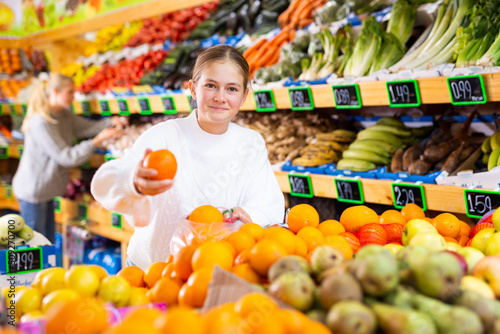 Portrait of happy teenage girl shopping in greengrocery store choosing ripe oranges..