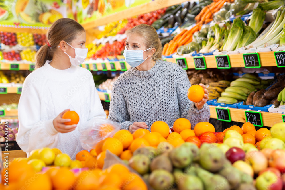 Portrait of teenage girl and her mother in protective mask who buying fresh oranges at grocery shop