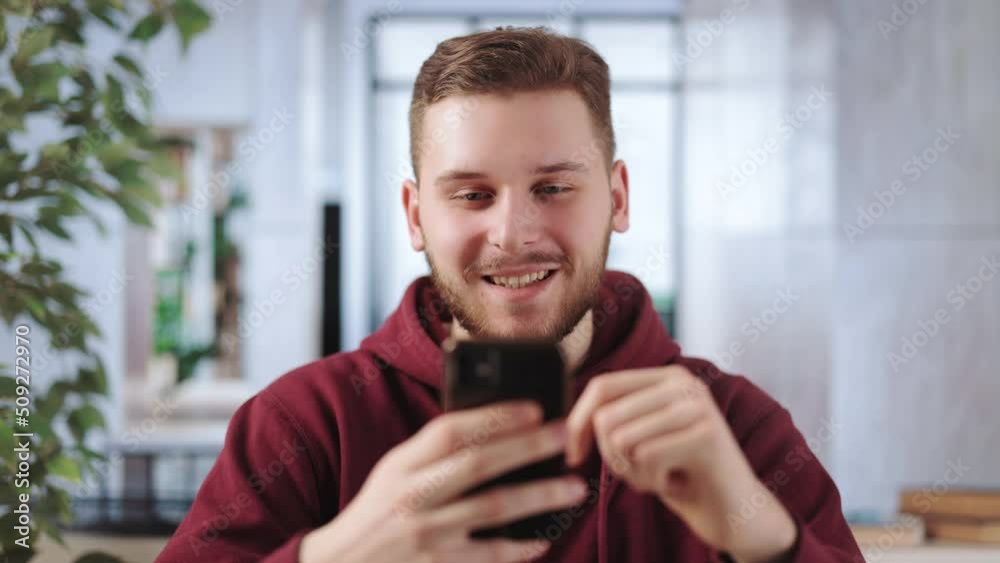 Positive young man scrolling news feed while resting from remote work in office. Caucasian male sitting at office desk with modern smartphone in hands. Positive young man scrolling news feed while resting from remote work in office. Caucasian male sitting at office desk with modern smartphone in hands.