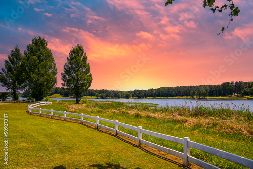 a gorgeous summer landscape at Lake Acworth with rippling blue lake water surrounded by lush green grass and trees with a white wooden fence and powerful clouds at sunset at Cauble Park photo