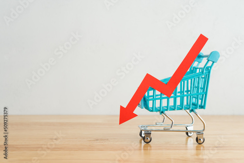 Shopping trolley with red chart falling down on wooden table background copy space. Economic recession crisis, core retail sales decrease, inflation or goods price up concept.