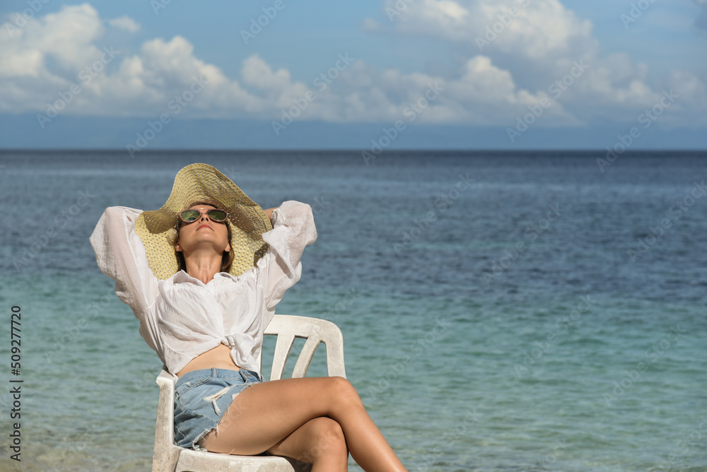 A lady in a straw hat, sunglasses, white shirt and denim shorts is sitting on a chair. A young girl is relaxing on the beach. Travel, happy holidays, tourism, Philippines, summer season, copy space