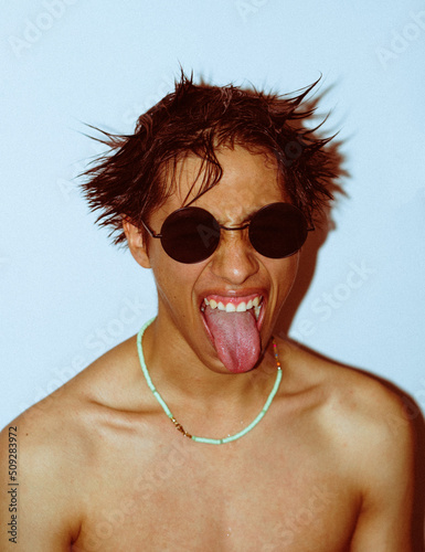 latin guy with black glasses sticks out his tongue at home with his shirt off, with retro 90s aesthetics photo taken with direct flash to the face, selective focus.