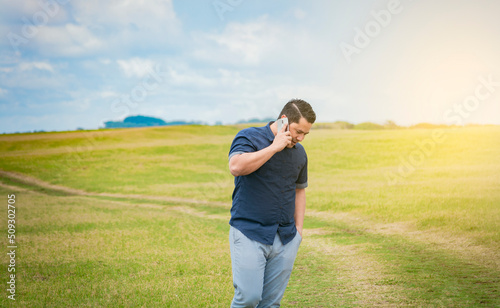 Person with his cell phone in the field talking on the phone, Man calling on the phone in the field, man on a road talking on the phone, young person talking on the phone in a field © IHERPHOTO