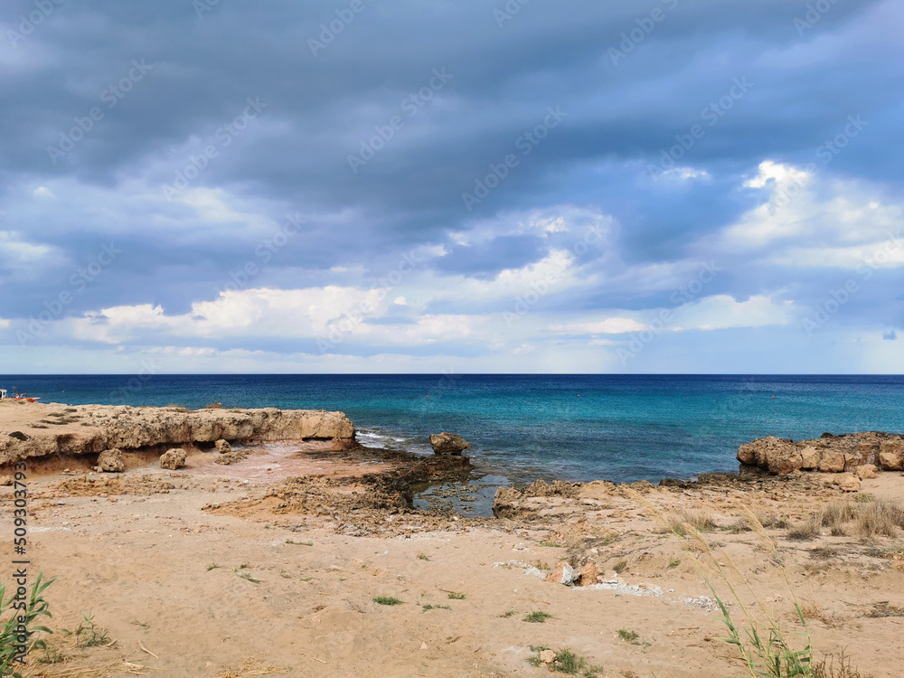 The sandy coast of the Mediterranean Sea, turning into stones from a long-hardened lava, azure water against a dramatic sky.