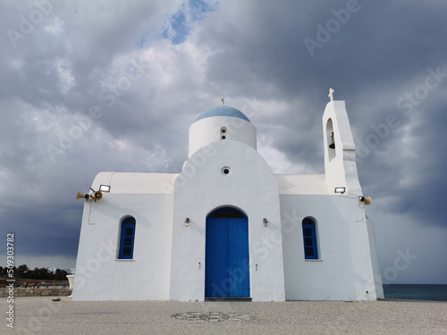 The Church of St. Nicholas the Wonderworker is white with a blue door against the backdrop of the Mediterranean Sea and the dramatic sky.