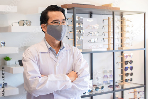 Masked optometrist over glasses display shelf holding hands crossed and look something in optical store, disease prevention concept