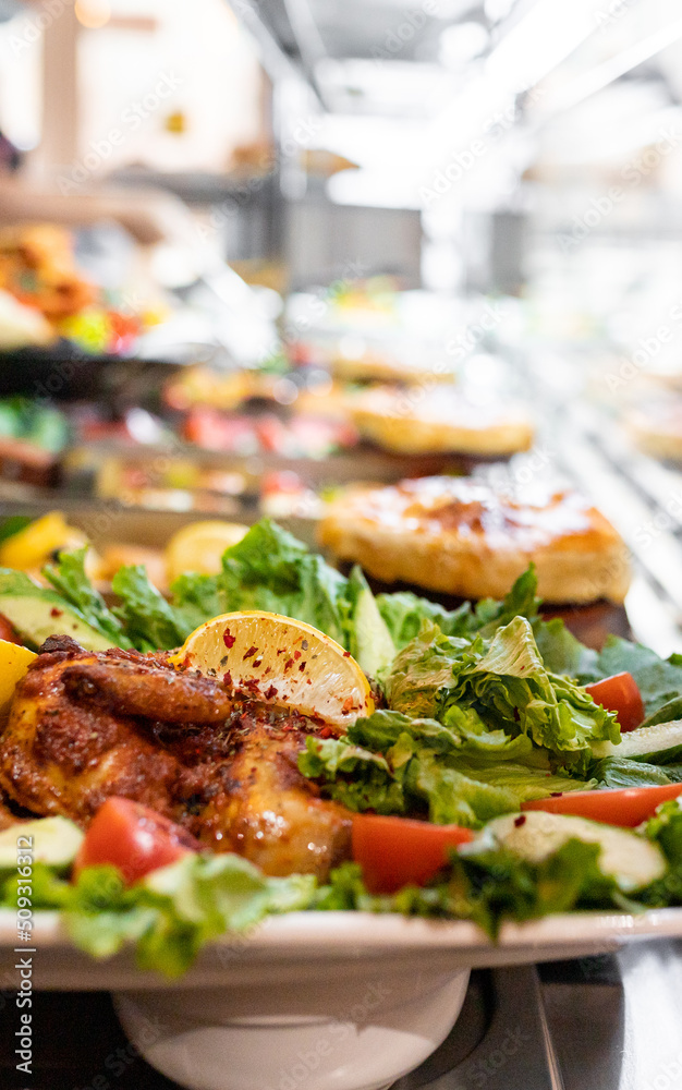 Tobacco chicken with lettuce, lemon and tomato among other out-of-focus dinner food. Selective focus, vertical copy space. Tobacco chicken with lettuce, lemon and tomato among other out-of-focus dinner food. Selective focus, vertical copy space.