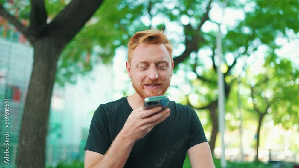 Cheerful red haired man recording voice message by phone while sitting on the bench in the park Cheerful red haired man recording voice message by phone while sitting on the bench in the park