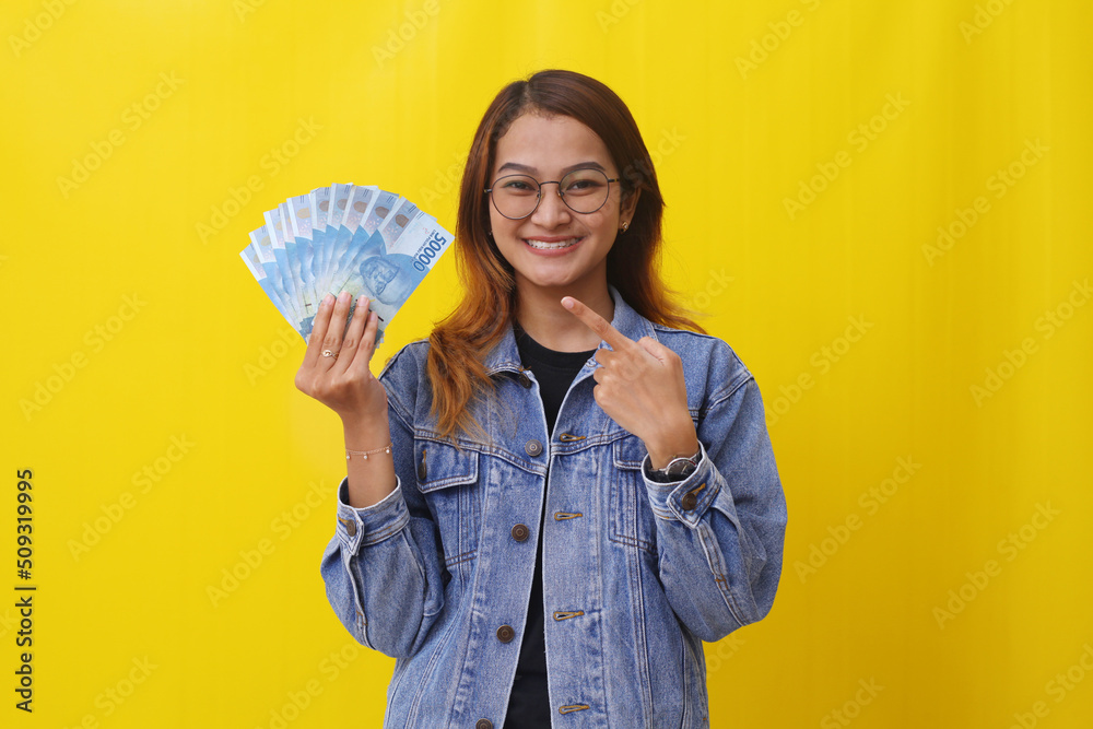 Happy young asian woman standing while holding and pointing an Indonesian money. Isolated on yellow Happy young asian woman standing while holding and pointing an Indonesian money. Isolated on yellow