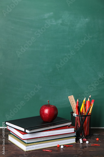 School stationery with books and apple on table near green chalkboard