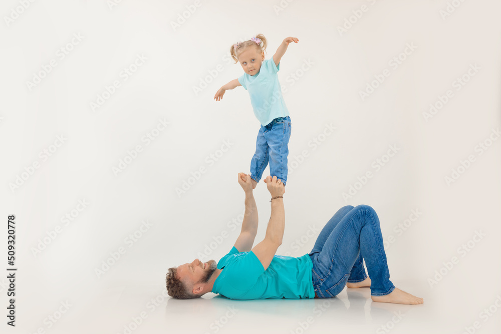 Athletic man with pumped up arms lying on back holding and lifting up with hands standing little girl, white background. Whole family workout, father and daughter. Sport and healthy lifestyle concept