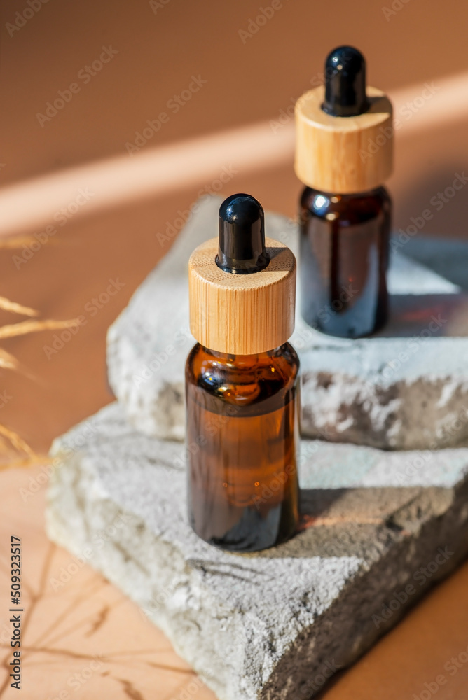 Amber glass droppers with bamboo lid and wild grass. Bottles on grey concrete podium for product presentation. Skincare cosmetic with wildflowers. Beauty concept