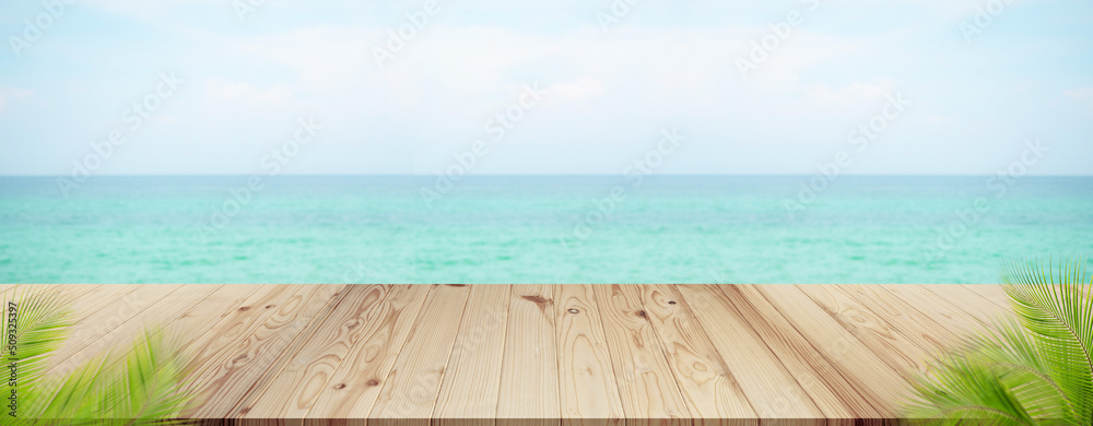 Fototapeta premium Palm leaves with wooden on sea.blank table on blue water shore and blue sky at coast. sunlight blur bokeh on ocean. mockup tropical seaside. Tourism relax vacation travel summer holidays.