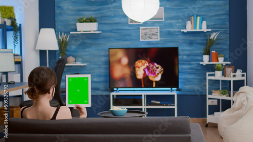 Girl holding vertical digital tablet with green screen in online conference or group video call in home living room. Young woman using touchscreen device with chroma key watching influencer vlog.