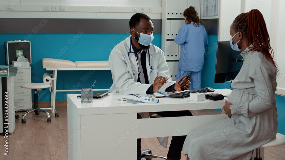 Medic and pregnant woman with face mask talking about pills treatment in bottle at checkup appointment. Patient with pregnancy belly receiving prescription medicine from doctor in cabinet.