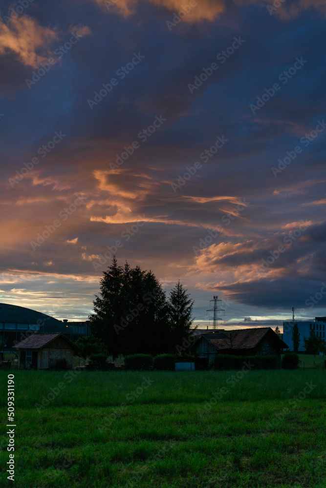 leuchtendes Abendrot nach dem Sonnenuntergang am Stadtrand von Dornbirn, Vorarlberg, Austria. tolle Stimmung mit roten und graublauen Wolken über den grünen Wiesen und Bäumen im Ried.