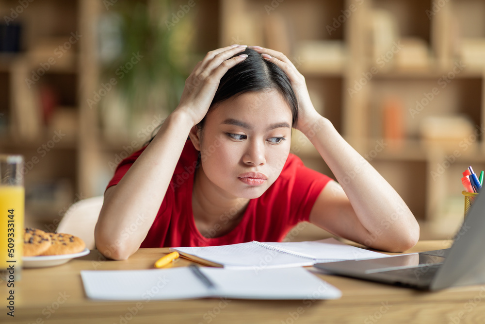 Tired upset teen chinese female hold head with hands, look at laptop in room interior Tired upset teen chinese female hold head with hands, look at laptop in room interior