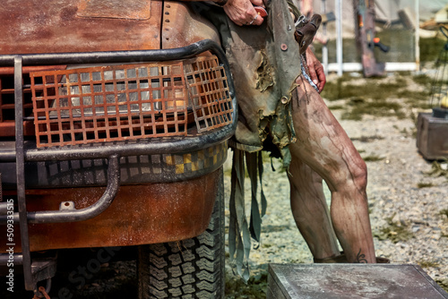 Completely rusted front part of suv with grills in front of headlight and dirty bare legs in background